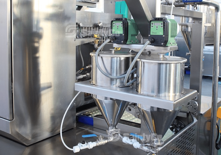 Close-up of industrial gummy manufacturing equipment with metal hoppers and attached piping. The machinery has green electronic components and wiring visible, looking clean and well-maintained in what appears to be a Gummygenix facility.