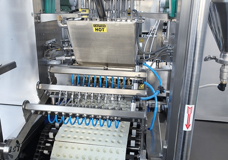 A SaintyCo gummy depositor with blue cables and multiple nozzles is actively dispensing a substance into white molds on a conveyor belt. A caution sign reading "HOT" is visible on a metal component. The surrounding area features stainless steel components and other industrial equipment.