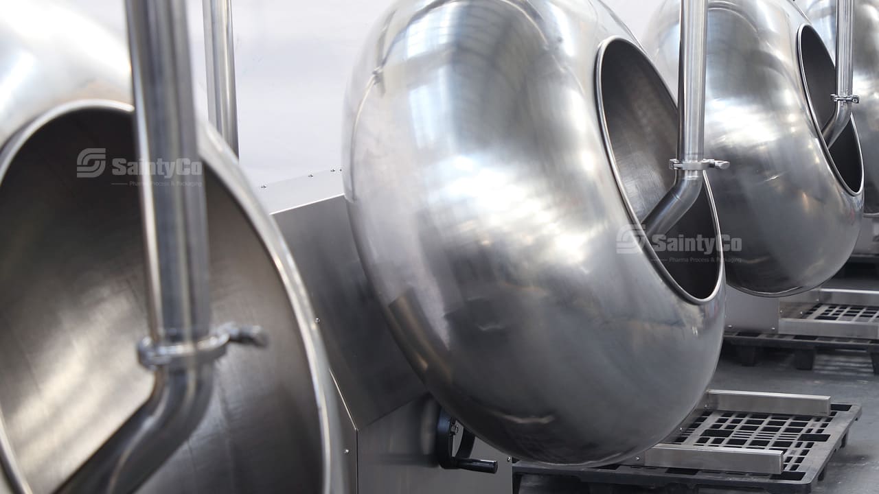 A row of large, shiny stainless steel industrial gummy making machines in a factory setting. The brand name "SaintyCo" is visible on the surface of the machines. They have cylindrical and bowl-like shapes, with various pipes and attachments extending from them.