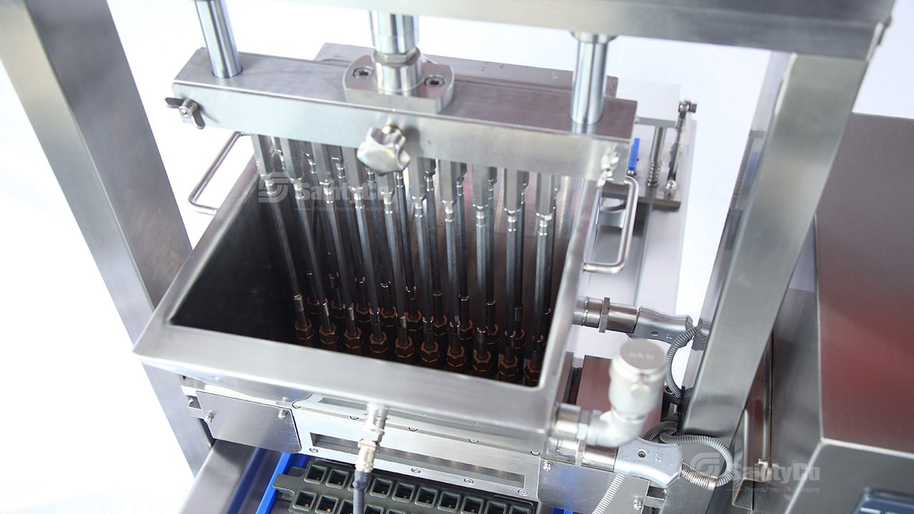 A close-up view of an industrial chocolate depositor machine in operation. Several nozzles dispense liquid chocolate into molds below, with a sleek stainless steel structure and precision components designed for efficient and uniform chocolate production, similar to gummy manufacturing equipment by SaintyCo.