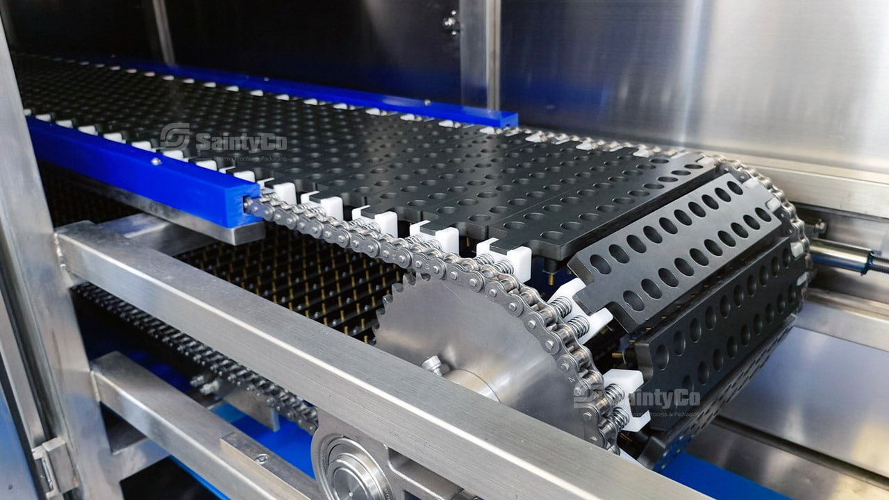 Close-up of a plastic chain conveyor in a gummy packaging machine by SaintyCo. The conveyor features a black, perforated belt and is housed within a metallic structure with blue accents. The name "SaintyCo" is visible on parts of the machine.