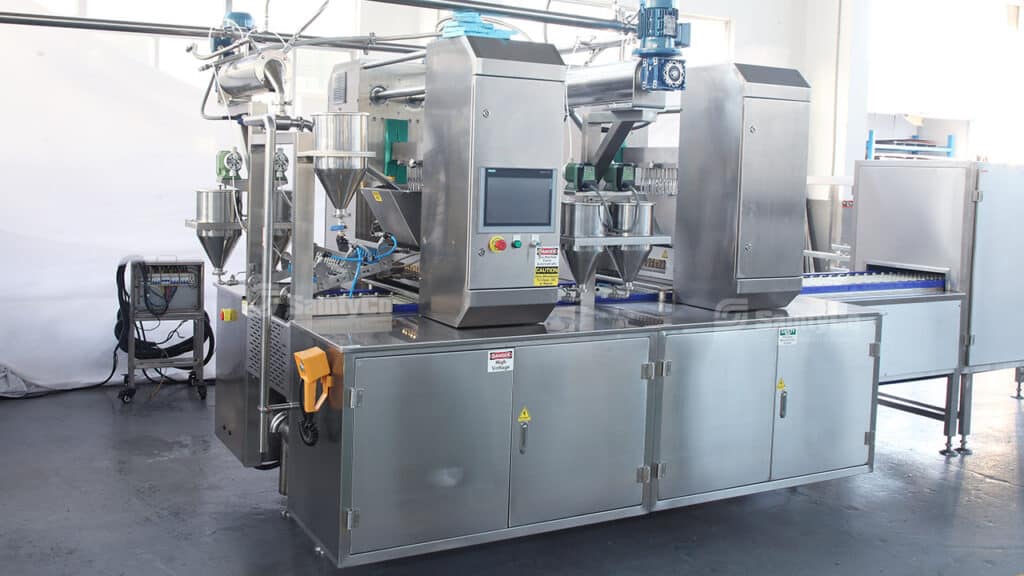 Large industrial gummy making machine with multiple stainless steel components, control panels, and a conveyor belt. It features various tubes and hoppers for material processing, with some safety labels and operational controls visible. The Gummygenix equipment is placed in a clean, bright factory setting.