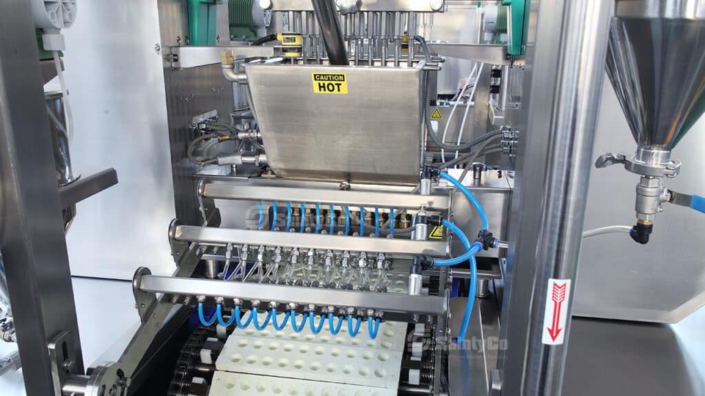 A close-up of a complex industrial machine, specifically SaintyCo's gummy manufacturing equipment, with numerous metal parts, blue wires, and a "CAUTION HOT" sign on a rectangular compartment. The machine appears to be involved in automated production, possibly dispensing or filling products on a conveyor belt.