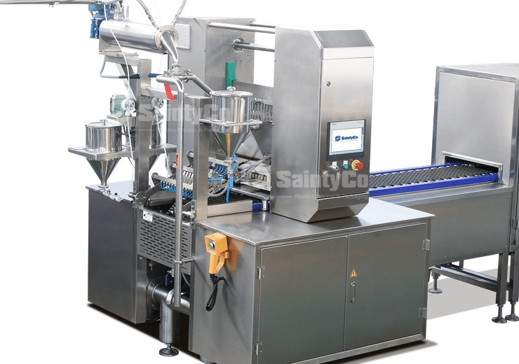 A stainless steel industrial food packaging machine with several interconnected components, including hoppers, conveyor belts, control panels, and tubing systems. The SaintyCo machine even includes a gummy depositor feature on the main control unit.