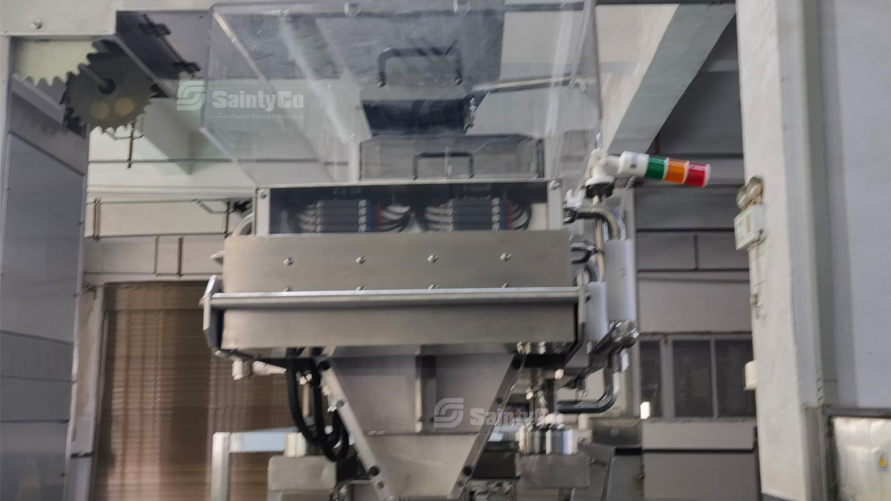 A close-up of a SaintyCo gummy making machine with a transparent cover, displaying internal components. The machine features a control panel with buttons and a multi-colored indicator light on the side. The background shows part of a factory.