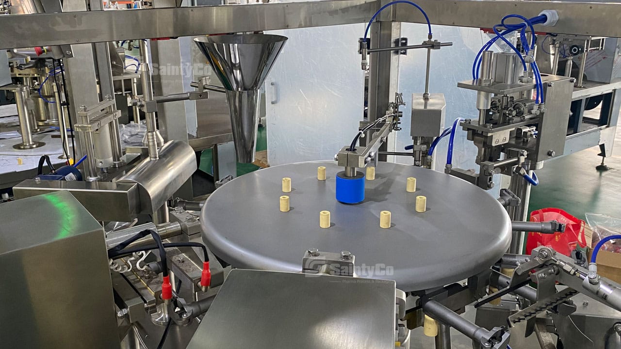 Industrial machinery with cylindrical components and a central rotating disc on which several yellow caps are placed. The background shows various metal parts and tubing, indicating a manufacturing or assembly process, typical of a SaintyCo gummy making machine.