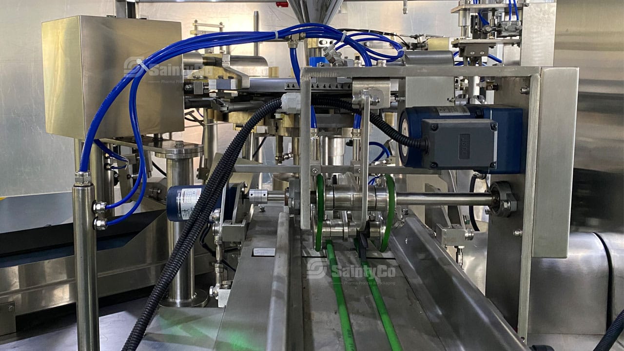 Close-up of industrial machinery with various metal components, blue tubing, and conveyor belts. The setup includes motors and mechanical parts, indicating a SaintyCo system for manufacturing or automated processing in a factory setting.