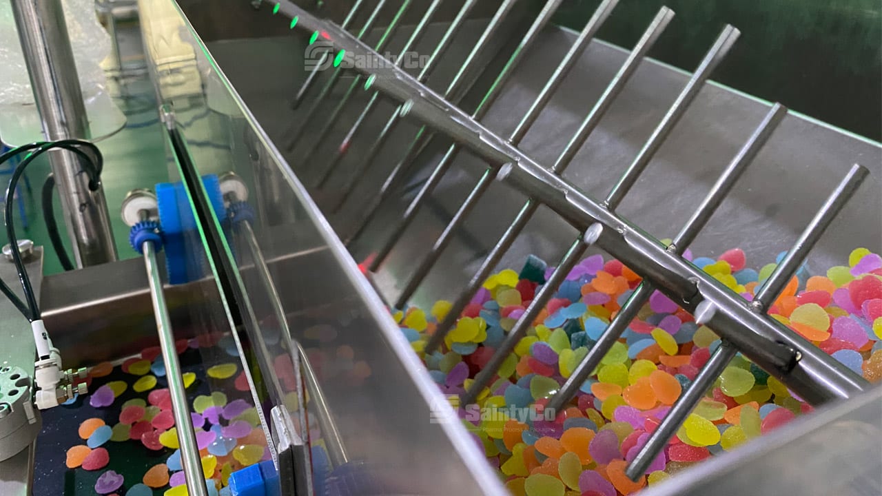 A close-up of a factory machine, possibly a SaintyCo gummy depositor, sorting colorful candies resembling gummies. The machine features a series of angled metal rods guiding the candies along the production line. Several vibrant, round candies can be seen accumulating at the bottom of the machine.