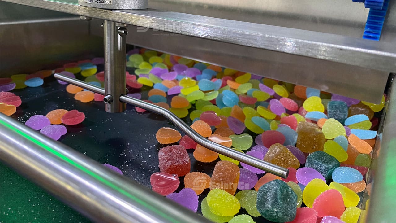 A gummy making machine in a candy factory is sorting a colorful array of jelly candies. The candies are in various shapes and bright colors, including red, pink, green, yellow, and blue. Metal rods are positioned above the candies as part of the sorting mechanism.