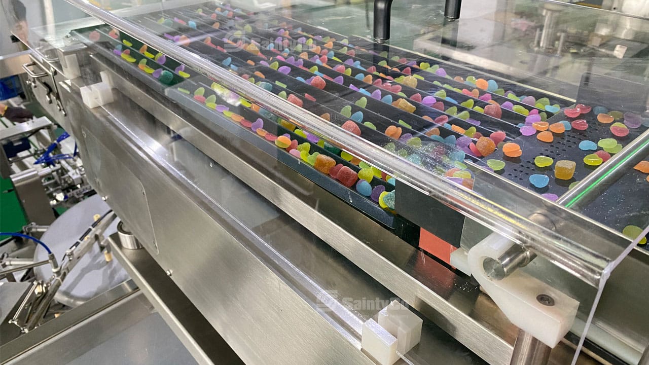 A close-up of a production line in a factory, with colorful gummy candies being processed and transported on a conveyor belt. The machinery, including a gummy making machine, is made of stainless steel with transparent covers protecting the candies. Various equipment parts are visible.