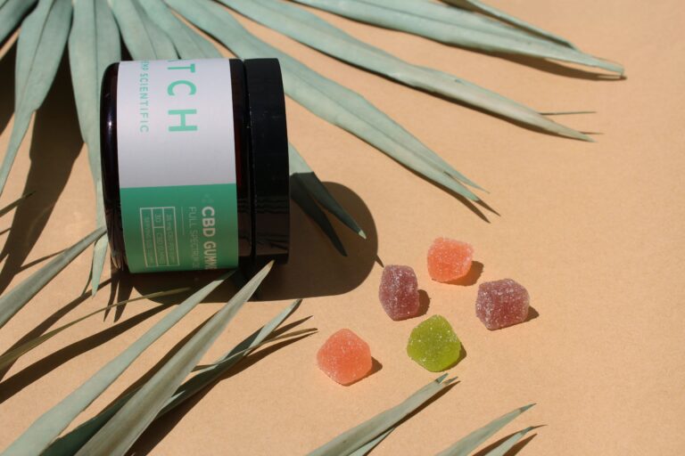 A container labeled “CBD Gummies” lies on its side, spilling out colorful, sugar-coated gummies in different shapes. The background features a peach-colored surface and striped green leaves, evoking a natural, tropical setting. It's as if the scene was crafted by a master gummy making machine.