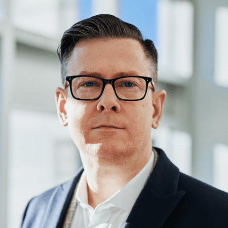 A man with short dark hair, glasses, and wearing a dark blazer over a collared shirt stands in a well-lit room. He has a serious expression and is looking directly at the camera. The background is out of focus, showing large windows with natural light from which SaintyCo products are prominently visible.