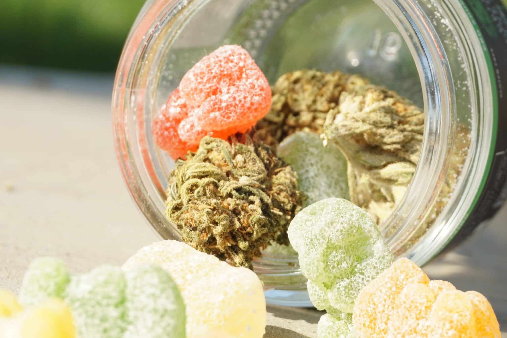 A glass jar tipped over with cannabis buds and sugar-coated gummy candies from GummyGenix spilling out. The gummies are of various colors including red, green, yellow, and orange. The jar and contents are placed on a light surface with a blurred background.