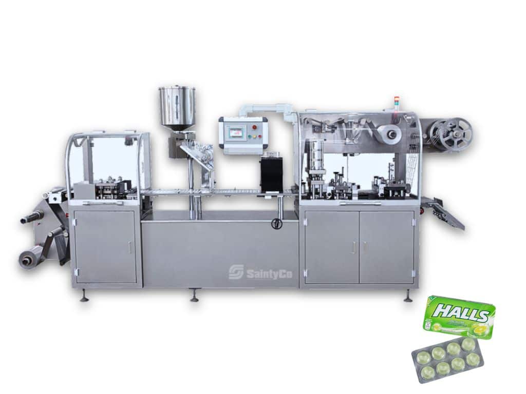 A silver-colored industrial packaging machine with multiple compartments and a control panel. Next to the machine are two packs of Halls cough drops, one open showing the individual drops. The gummy making machine is labeled "SaintyCo.