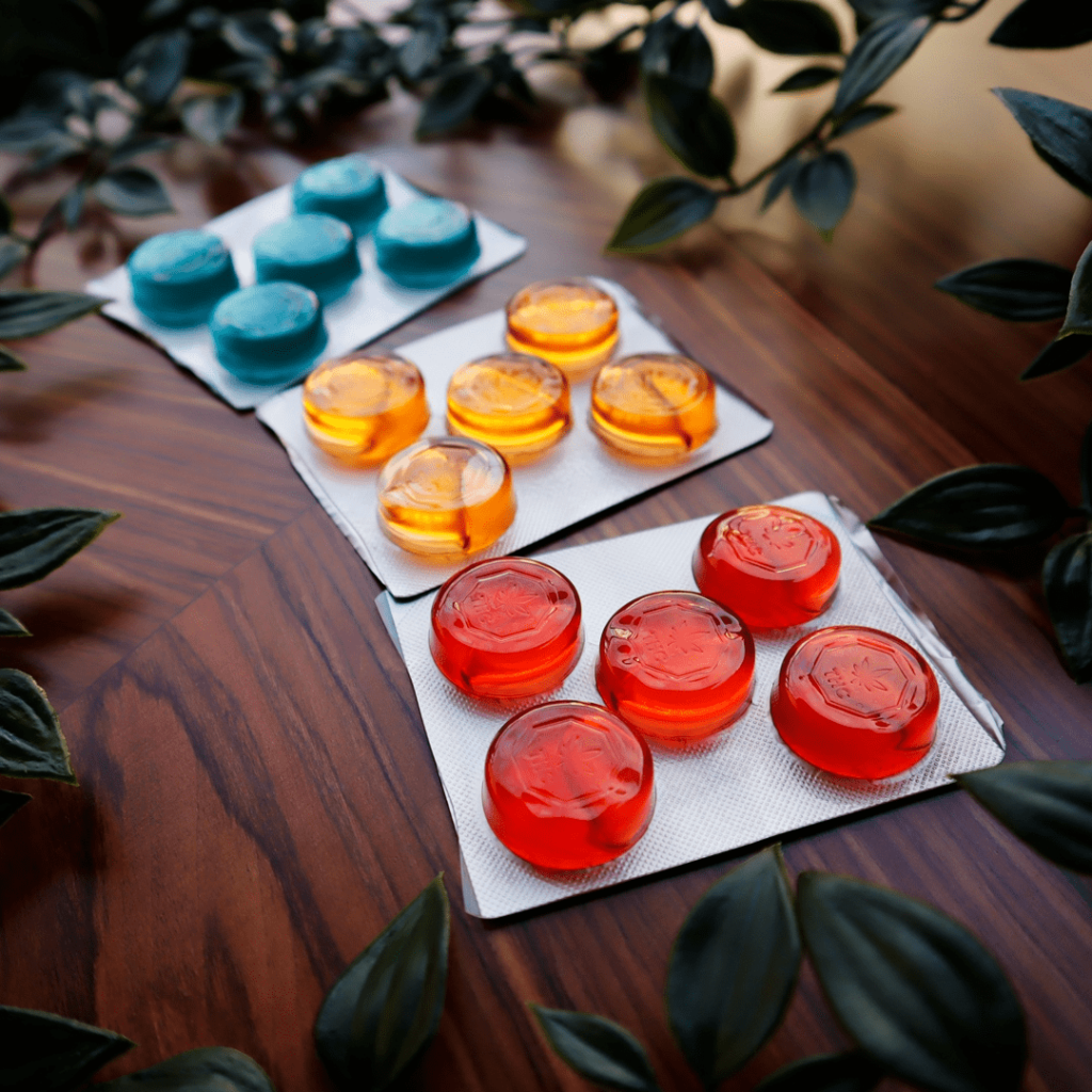 Three blister packs containing round, colored gummies are laid out on a wooden surface surrounded by green leaves. The gummies' colors are blue, orange, and red, with each pack holding three gummies. The setting seems natural and inviting, showcasing the vibrant results from a state-of-the-art gummy depositor.