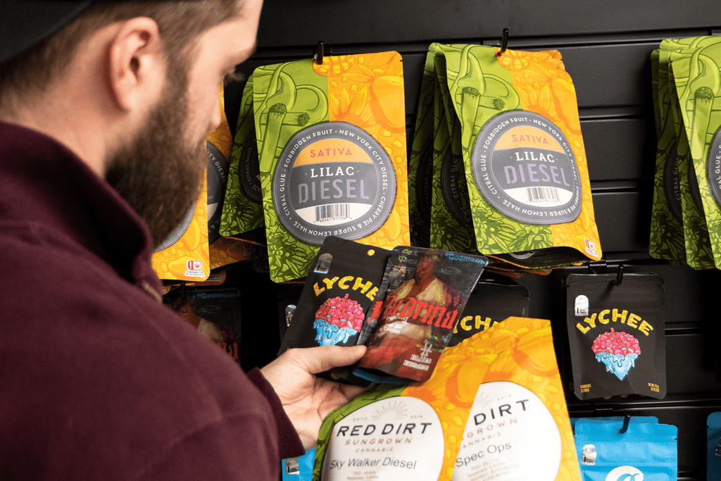 A person in a maroon jacket and black cap looks at a product in their hand while standing in front of a display of cannabis products, likely considering how gummy manufacturing equipment from brands like SaintyCo or Gummygenix could enhance production. The packaging on the wall includes labels like "Lilac Diesel," "Lychee," and "Red Dirt.