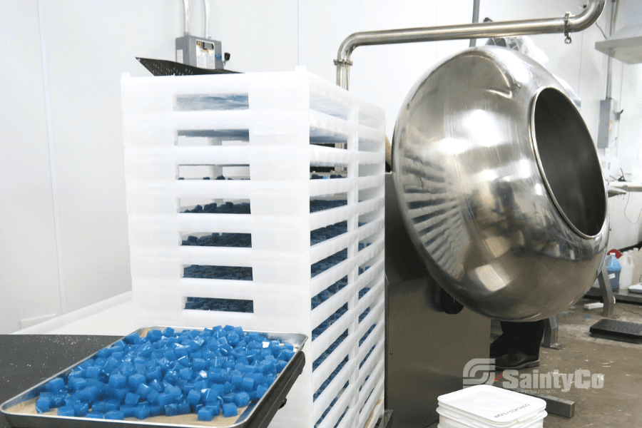 A stainless steel gummy depositor in a factory setting is positioned next to a tall stack of white trays filled with numerous blue cube-shaped items. A tray with additional blue cubes is placed on a table in the foreground. The logo "SaintyCo" is visible in the image, indicating it's part of their gummy manufacturing equipment.