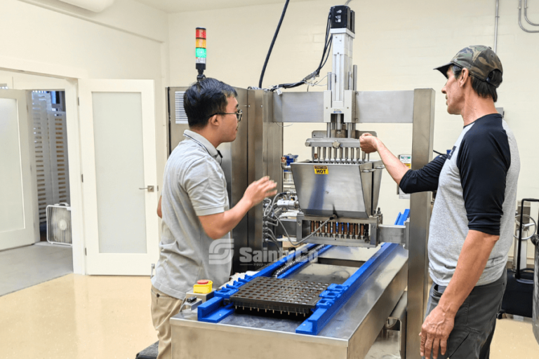 Two men are working with a piece of industrial machinery inside a workshop. Engaging with SaintyCo gummy manufacturing equipment, they focus on the complex machine’s levers and components. One man, wearing glasses, appears to be explaining something related to the Gummygenix process while the other listens attentively.