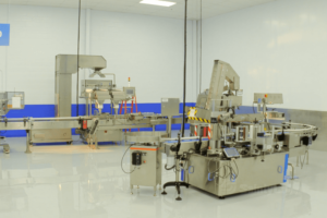 A clean, modern industrial facility with automated machinery, featuring conveyer belts and stainless steel equipment from SaintyCo. The room has bright lighting, white walls, and a shiny, polished floor. There are control panels and indicator lights on the gummy making machine.