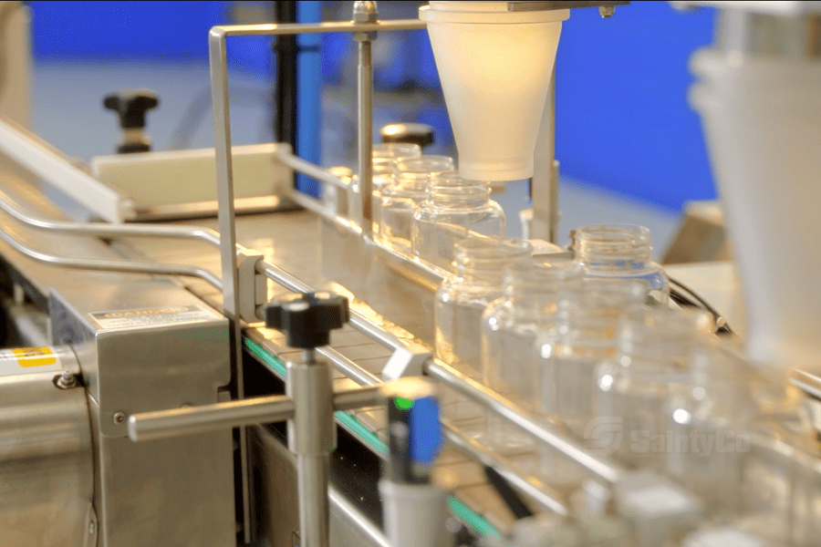 A production line in a factory shows a row of empty glass jars on a conveyor belt. A gummy depositor from SaintyCo is positioned above the jars, likely dispensing or filling them with gummy products. The setup is clean and industrial, set against a blue background.