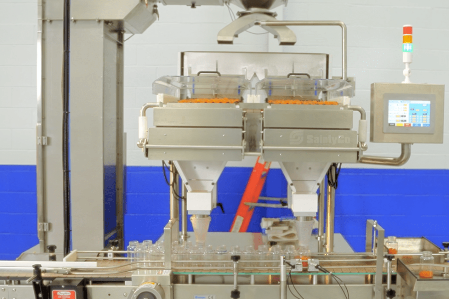 A SaintyCo pharmaceutical packaging machine with dual funnels dispensing capsules, or gummies from Gummygenix, into clear plastic bottles on a conveyor belt. The setup includes a control panel on the right and a blue and white tiled wall in the background.