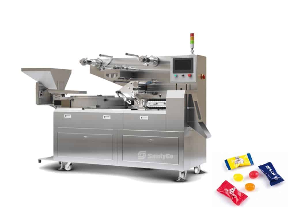 A stainless steel candy wrapping machine, often used alongside gummy manufacturing equipment, features a control panel, various rollers, and a conveyor system on the left. On the right side, a few wrapped candies in colored packaging are displayed.