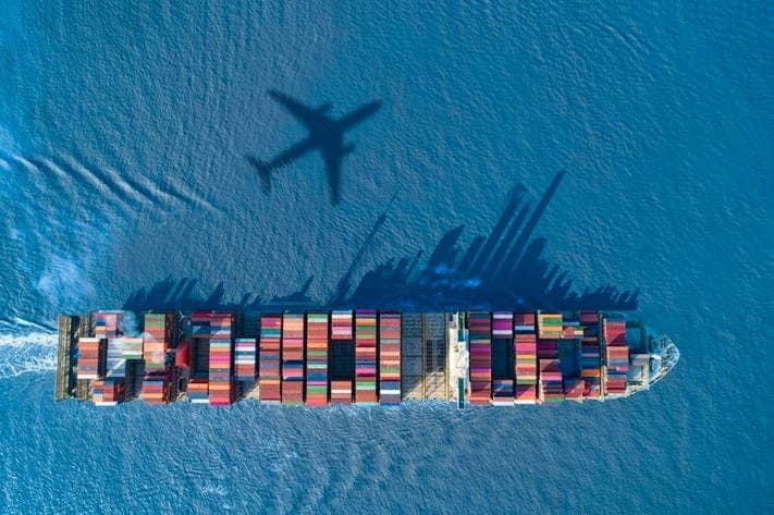 Aerial view of a large cargo ship laden with colorful shipping containers, sailing through deep blue water. The shadow of an airplane is prominently cast on the ocean surface beside the ship, almost reflecting the precision found in a SaintyCo gummy making machine.