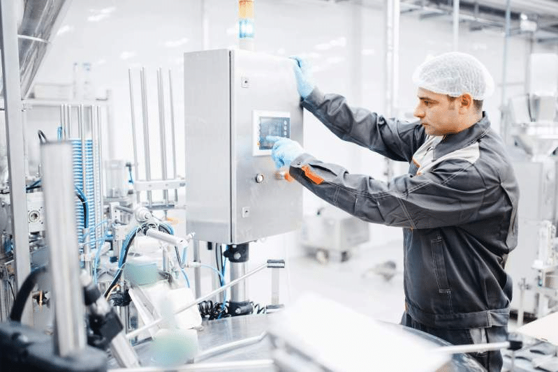 A factory worker wearing a hairnet, gloves, and a protective uniform operates a control panel on an industrial gummy depositor in a clean, modern manufacturing facility. The worker's focus is on the touchscreen interface of the machinery.