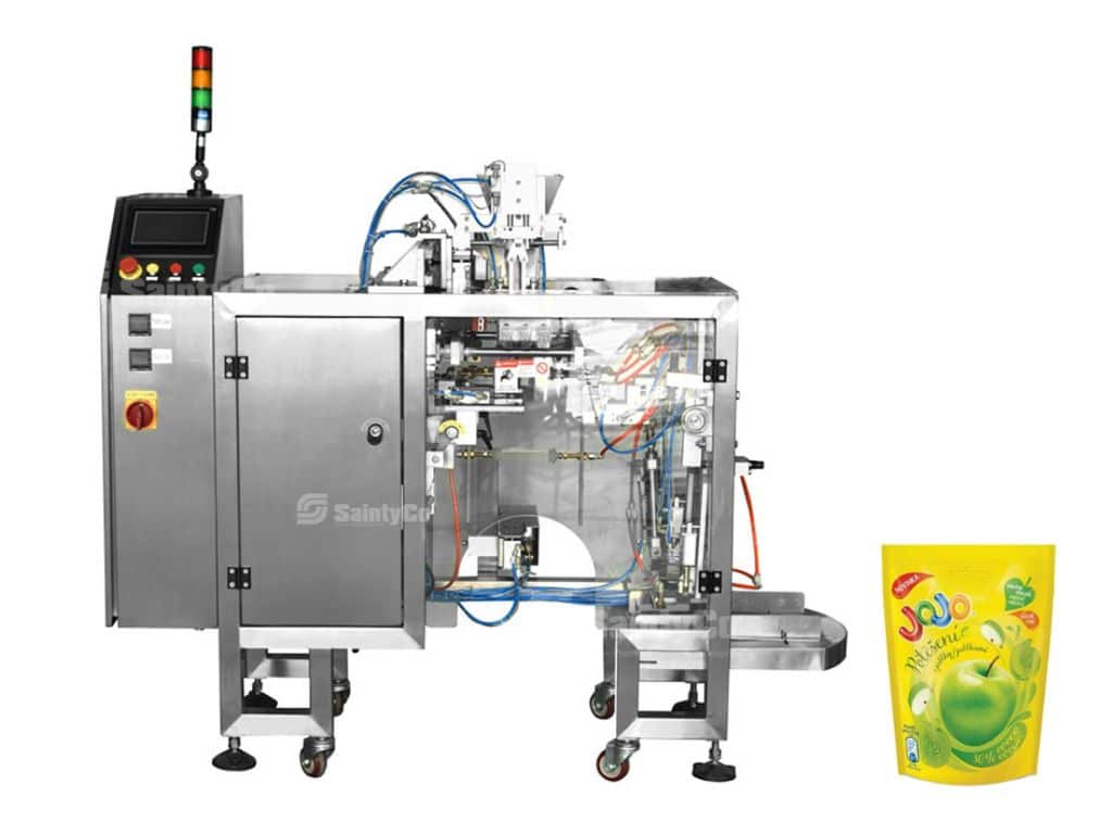 A stainless steel packaging machine from SaintyCo stands on wheels, equipped with various wires and tubes, a control panel with a red emergency stop button, and a multi-color signal light. Next to it is a brightly colored, sealed pouch of green apple juice labeled "Jojo".