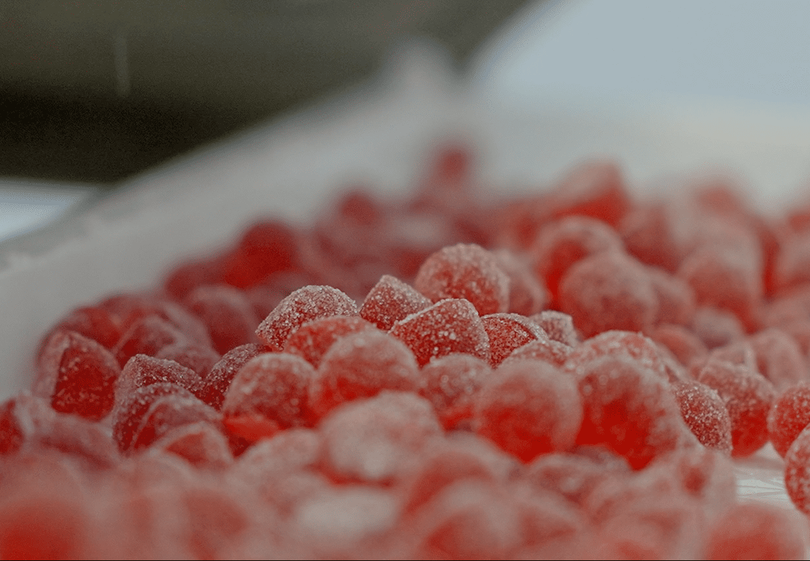 Close-up of a pile of small, round red gelatin candies coated with sugar on a white surface. The candies appear shiny and slightly translucent, with a soft, chewy texture suggested by their appearance—perfectly crafted using Gummygenix gummy manufacturing equipment.