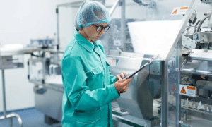 A person wearing glasses, a hairnet, and a teal lab coat is holding a clipboard while inspecting a gummy making machine in a laboratory or manufacturing setting. The equipment has caution labels, and the environment appears clean and sanitary.