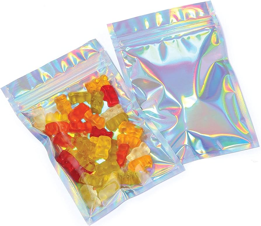 Two shiny, iridescent resealable bags are shown, one open and filled with various colored gummy bears from a SaintyCo gummy depositor, and the other sealed and empty. The glossy surface of the bags reflects a spectrum of colors.