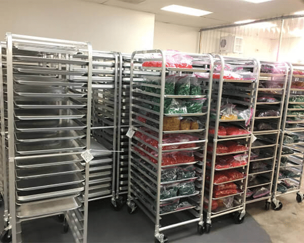 Multiple metal racks on wheels are shown in a room. Some racks are empty, while others hold bags of colorful, packaged gummy items. The room appears to be industrial, possibly a kitchen or storage area featuring a gummy depositor from SaintyCo. The floor is gray and the walls are plain and light-colored.