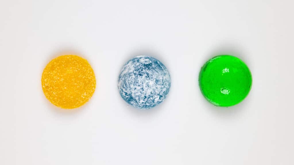 A yellow, blue-speckled, and green circular object are arranged in a horizontal row on a plain white background, resembling the output of a gummy making machine. The objects appear to be small, smooth, and glossy.