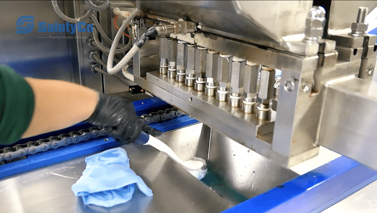 A person wearing a black glove uses a brush to clean the inside of a stainless steel gummy making machine with multiple nozzles. A blue cleaning cloth is placed next to the person. The machine has the "SaintyCo Pharma Process" logo on the left side.