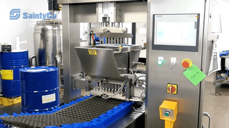 A manufacturing machine with a digital control panel displaying various buttons and settings. The gummy depositor is processing blue barrels on a conveyor system, with one barrel in position under the metallic apparatus. The logo "SaintyCo" is visible on the left.