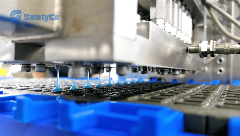 Close-up of a tablet press machine with a blue logo "SaintyCo" in the top left corner. The machine, resembling advanced gummy manufacturing equipment, is pressing and forming tablets, with multiple nozzles dispensing powder into molds on a conveyor belt system.