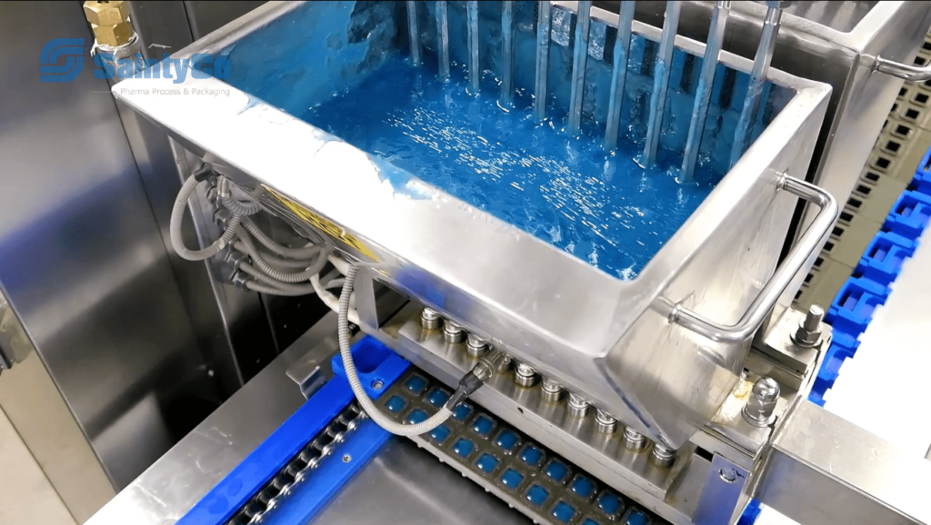 Factory machinery with a stainless steel container filled with blue liquid operates as part of a gummy making machine. Multiple pipes are submerged in the liquid, and below the container, a conveyor system with a blue plastic tray is visible. The equipment appears to be used for an industrial process.