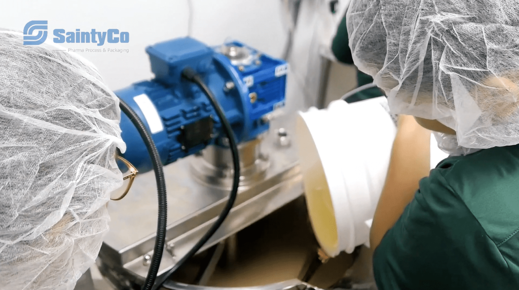 Two workers in hairnets and green uniforms are handling a white container, pouring its contents into a stainless steel gummy depositor of GummyGenix by SaintyCo. The machine has a blue motor and the SaintyCo logo is visible in the top left corner, indicating a pharmaceutical or gummy manufacturing equipment setting.