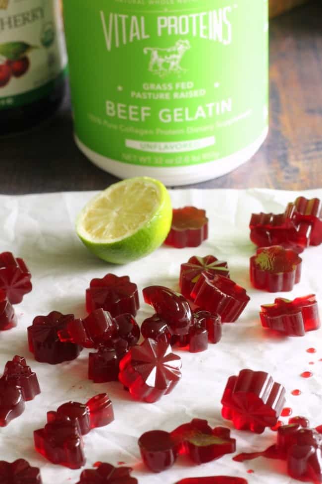 Assorted flower-shaped red gummies made using GummyGenix gummy manufacturing equipment are scattered on a white parchment paper. A halved lime and a green container labeled "Vital Proteins Beef Gelatin" from SaintyCo are in the background.