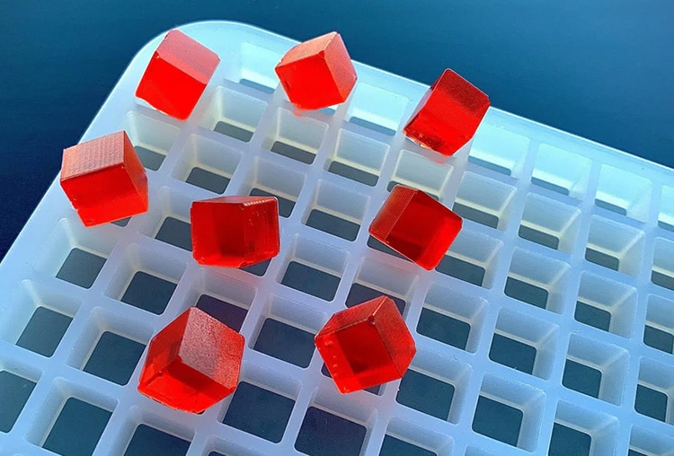 Red cube-shaped gelatin pieces, resembling gummy treats, are arranged on a white plastic grid tray against a dark blue background. The transparent red cubes are glossy and evenly spaced across the tray's square holes, as if meticulously crafted by a professional gummy making machine.