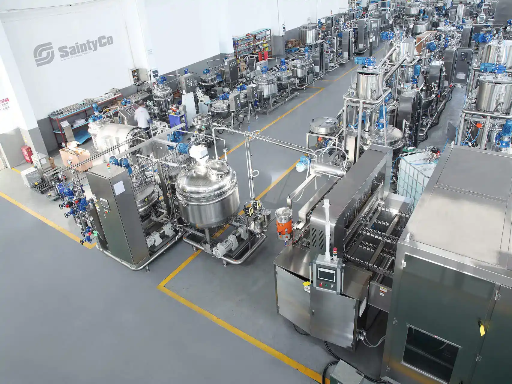 The image shows a modern, organized manufacturing facility. It features various industrial machines, large stainless steel tanks, and equipment arranged in rows. Several workers in white lab coats are seen operating the machinery. The floor is clean with marked pathways.