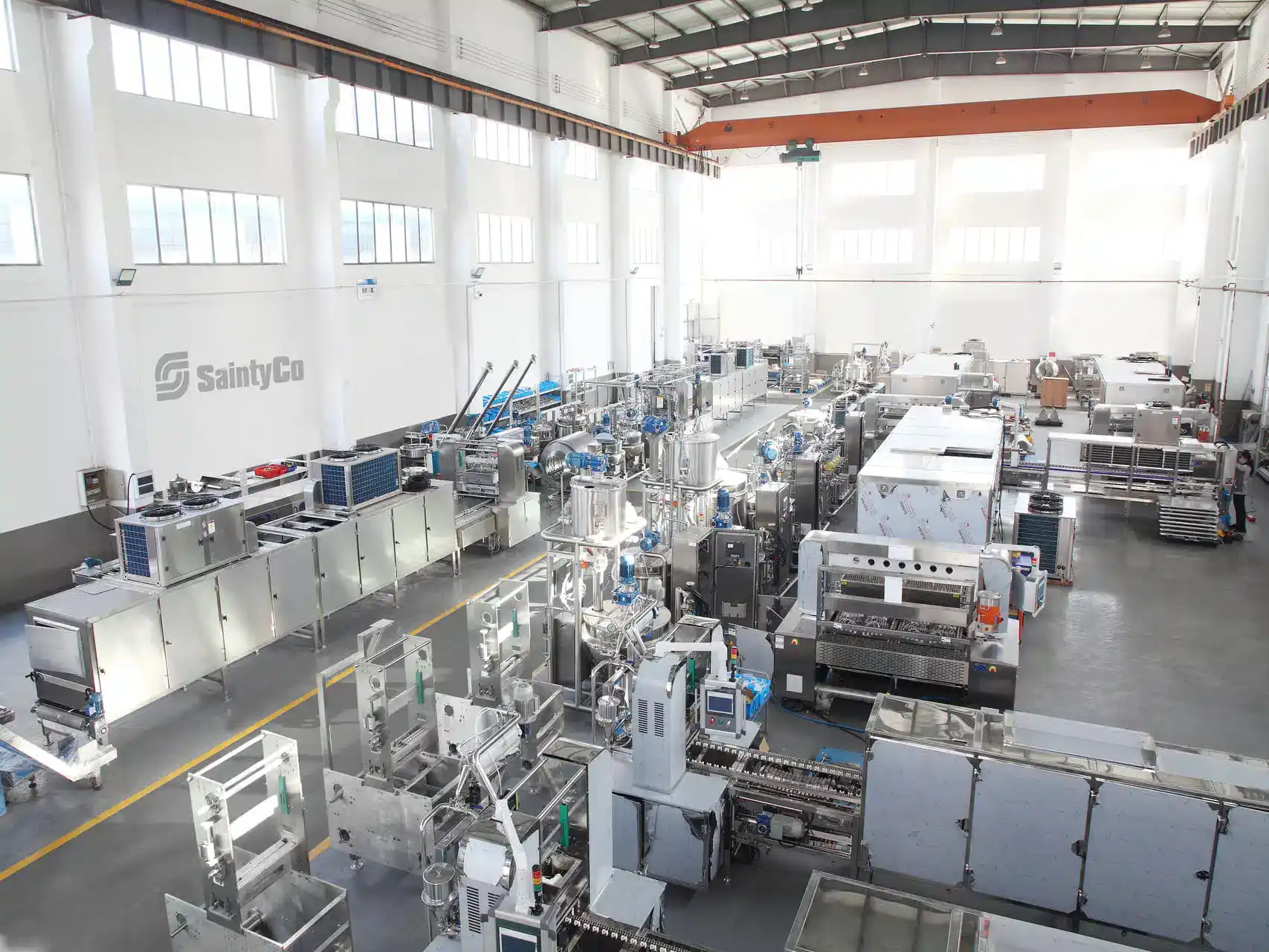 An expansive industrial facility featuring numerous large machines, including a gummy depositor, arranged in a spacious, well-lit area with high ceilings. The SaintyCo logo is visible on one of the walls. The machinery appears to be related to manufacturing or processing.