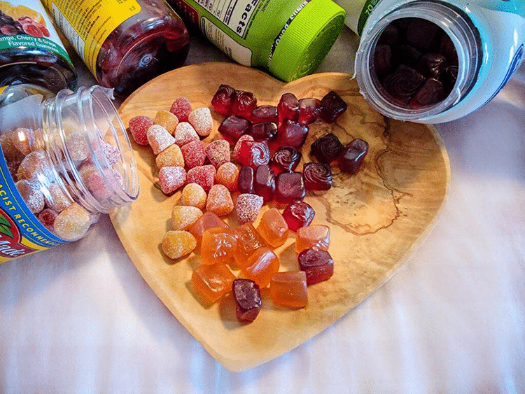 A variety of GummyGenix by SaintyCo supplements are scattered on a heart-shaped wooden plate. Several open bottles surround the plate, each containing differently shaped and colored gummies produced by advanced gummy making machines. Some gummies are translucent, while others are coated in sugar.