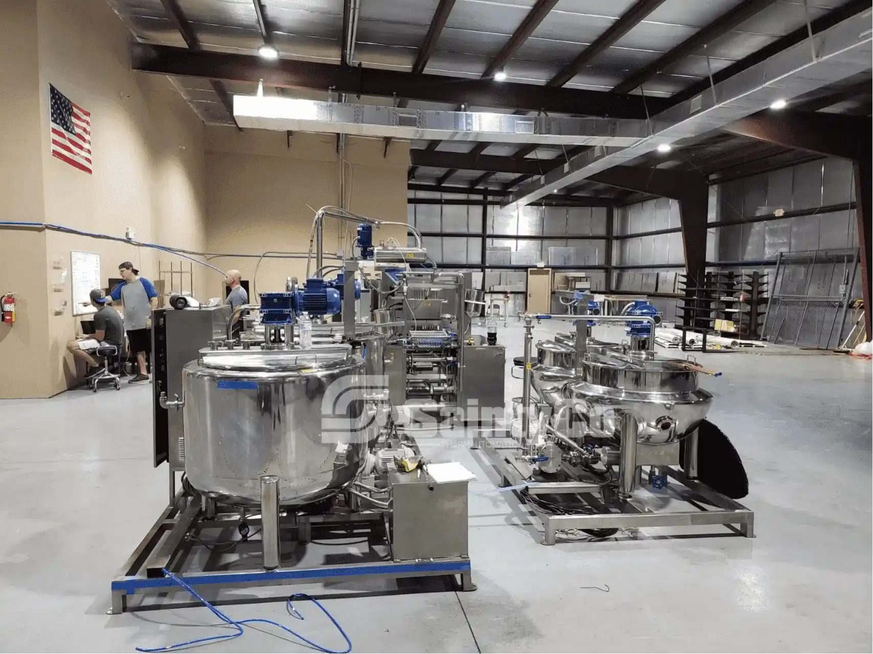 A modern industrial facility with large, stainless steel machinery and equipment for manufacturing or processing gummies. Workers are seen in the background at their workstations operating a state-of-the-art gummy depositor. An American flag hangs on the wall. The space has high ceilings and is well-lit.