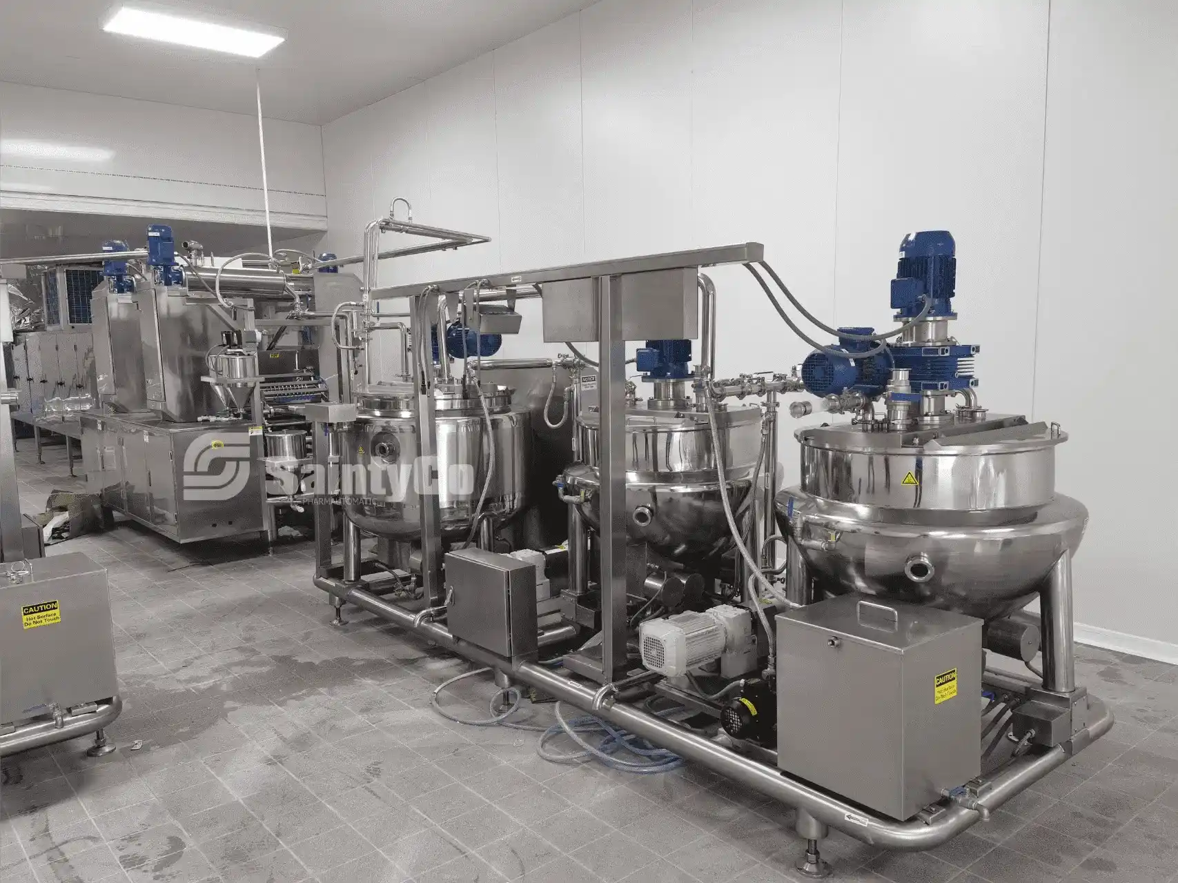 A clean and modern industrial kitchen featuring SaintyCo's high-end gummy manufacturing equipment, including large stainless steel mixing and processing machines, several tanks, and pipes. The sterile, white-walled environment indicates it's designed for food or pharmaceutical production.