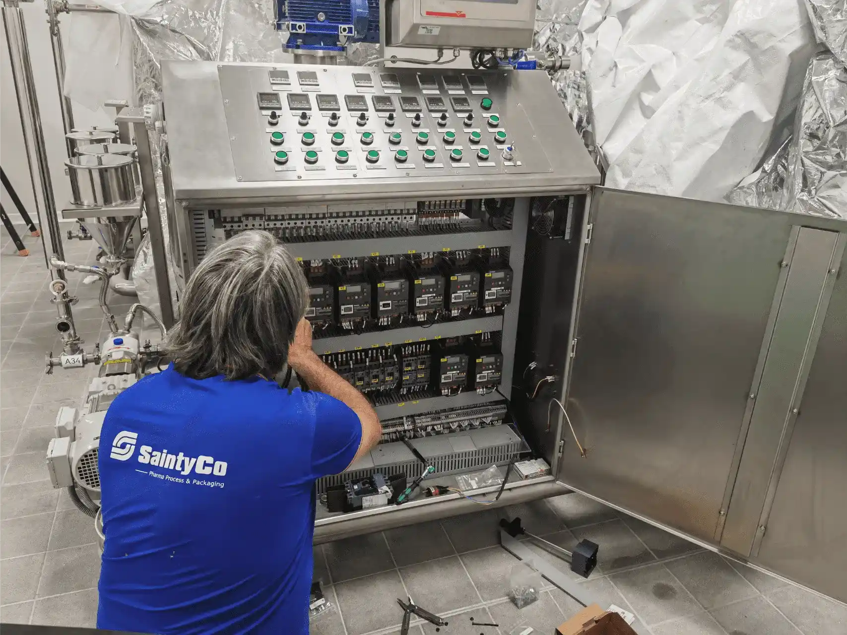 A technician in a blue "SaintyCo" shirt is working on an open electrical panel with various switches, buttons, and wiring. The panel is part of industrial machinery in a modern, clean facility. The technician is adjusting components inside the panel.