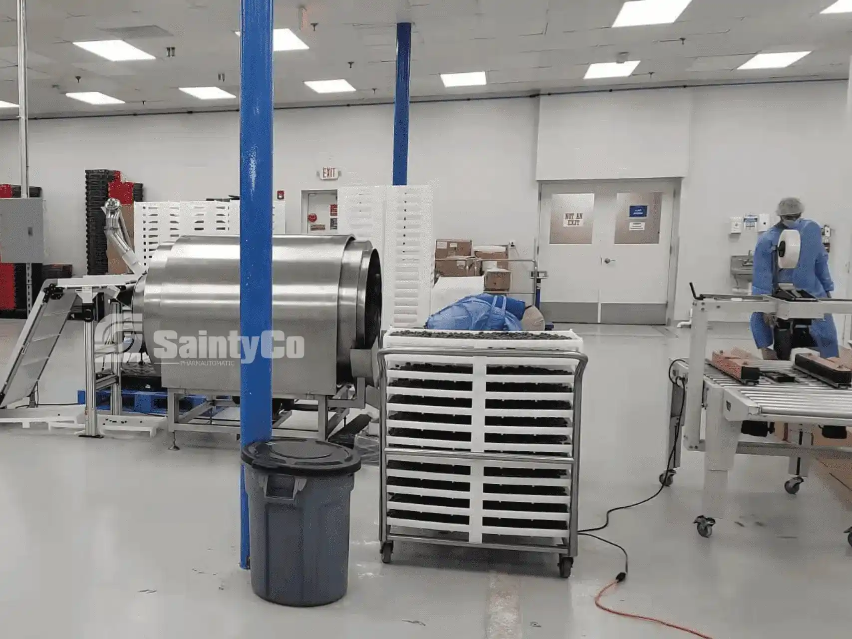 A clean, well-lit pharmaceutical manufacturing room with stainless steel equipment and stacked trays. Two workers in blue protective gear and hairnets are seen working with gummy manufacturing equipment. The area appears organized and sterile, featuring blue support columns.