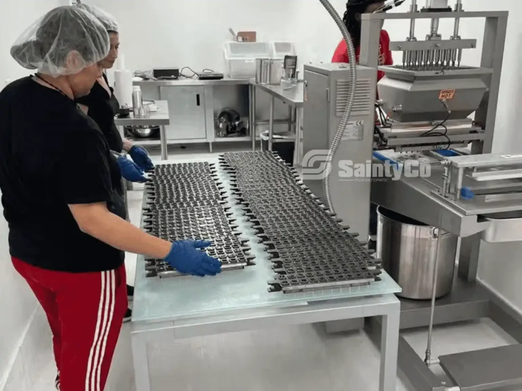 Two individuals in protective gear and gloves operate a SaintyCo gummy depositor in a sterile laboratory setting. They meticulously manage rows of molds or trays on a table, indicative of an industrial process related to food production, particularly gummies.