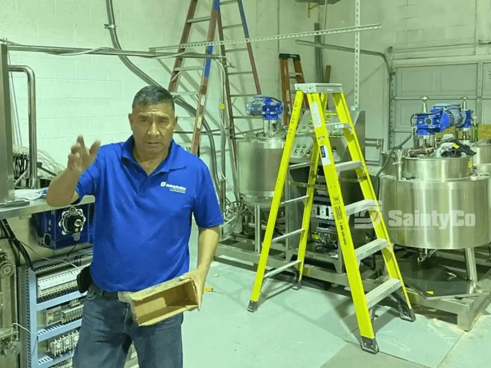 A man in a blue polo shirt stands in a room with industrial equipment. He holds a small wooden crate in one hand and gestures with the other. Behind him are a ladder and large metal tanks, alongside a state-of-the-art gummy depositor. The room has pipes and wires along the walls, with a "SaintyCo" logo visible.